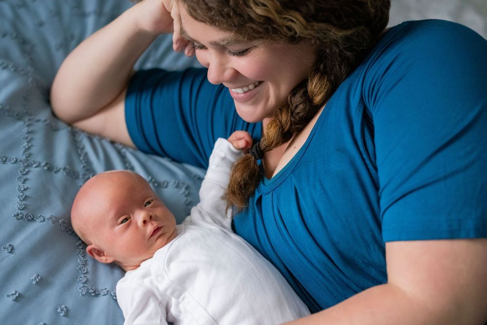 Smiling mother in blue shirt lying on bed gazing lovingly at her newborn baby.
