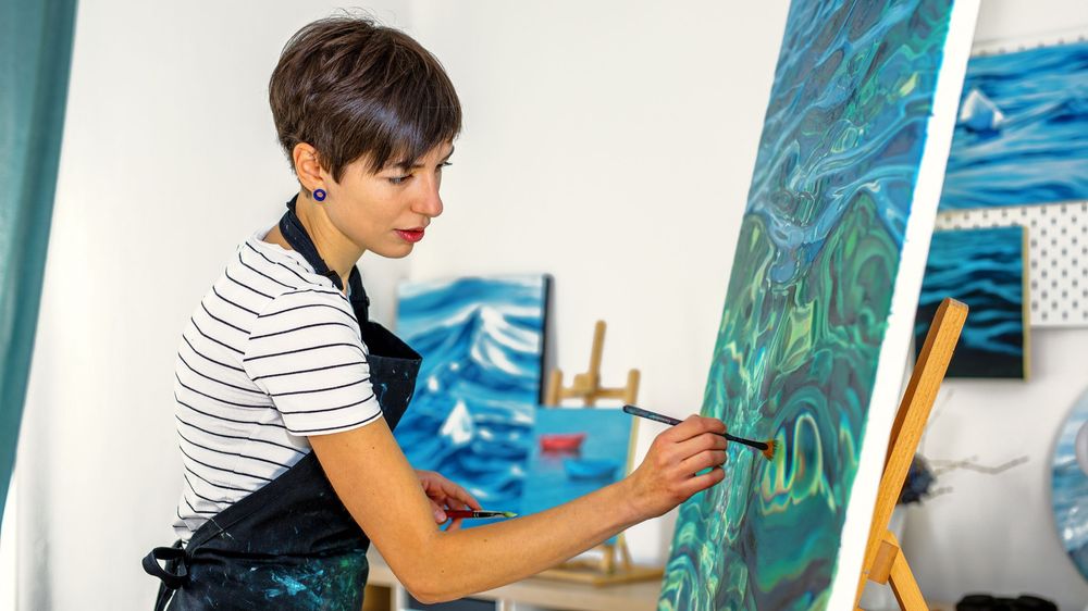 Artist in striped shirt working on vibrant ocean-themed painting at easel in art studio.