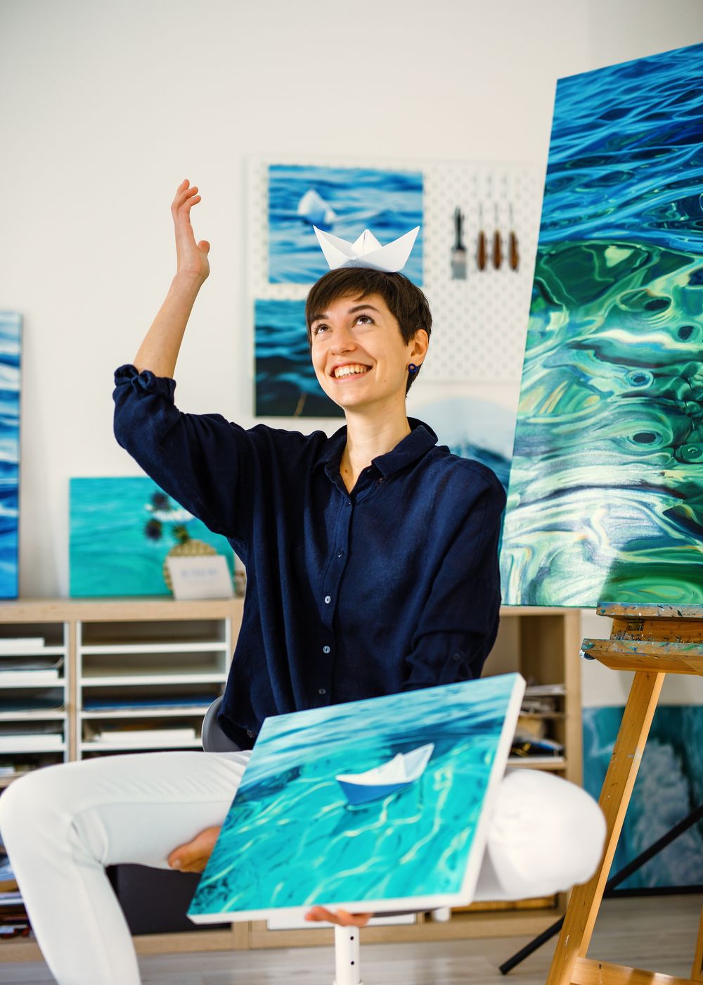 Artist in navy shirt sits among vibrant ocean-themed artwork in bright studio space with raised arm.