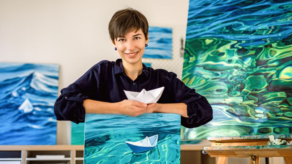 Artist holding paper boat against backdrop of ocean-themed artwork in studio setting.