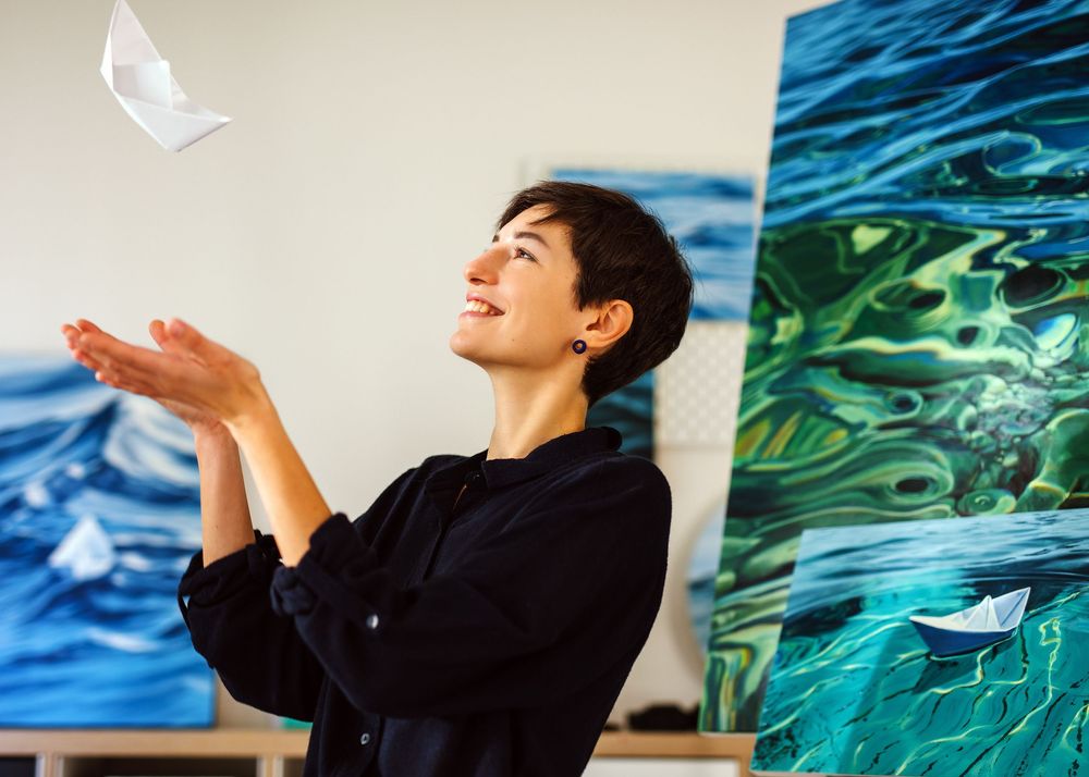 Artist playfully tossing paper boat in studio with ocean paintings displayed in background.