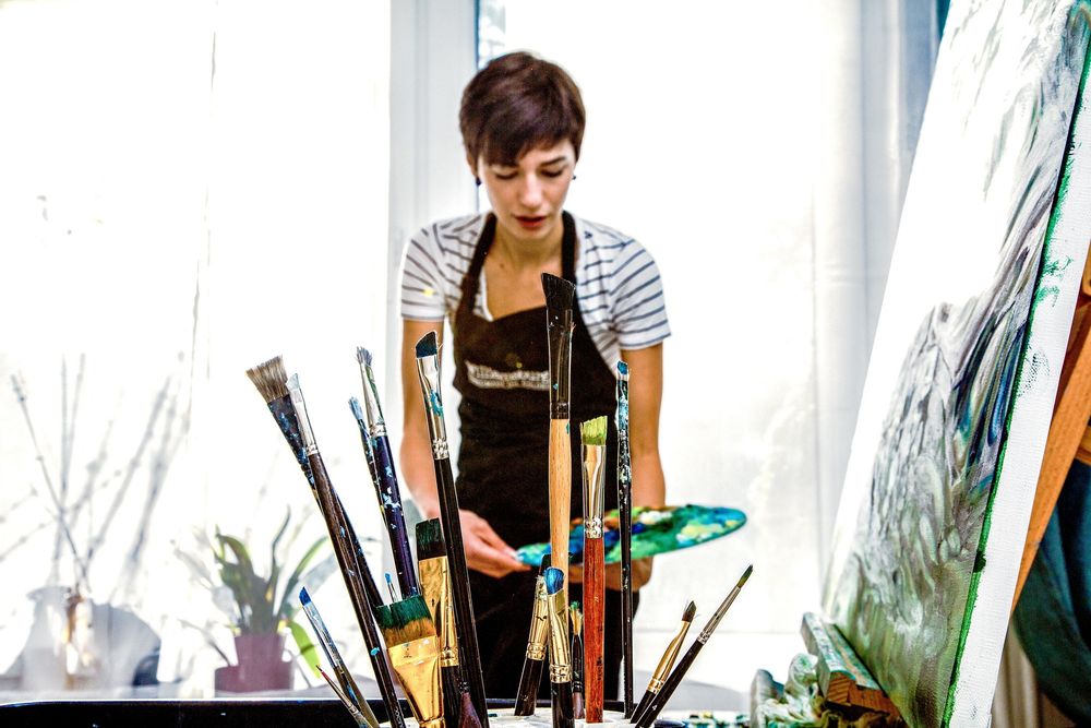 Artist in striped shirt stands in studio surrounded by paintbrushes and ocean-inspired artwork.