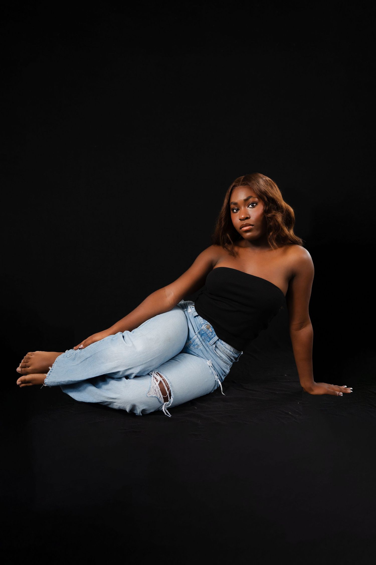 Young Black woman poses on dark floor in black strapless top and ripped light blue jeans, looking confidently at camera.