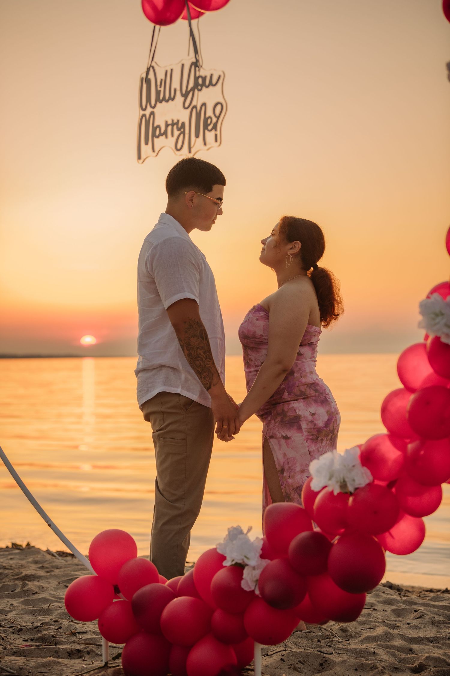 Couple holding hands during a romantic beach sunset proposal with red balloons and a Will You Marry Me sign.