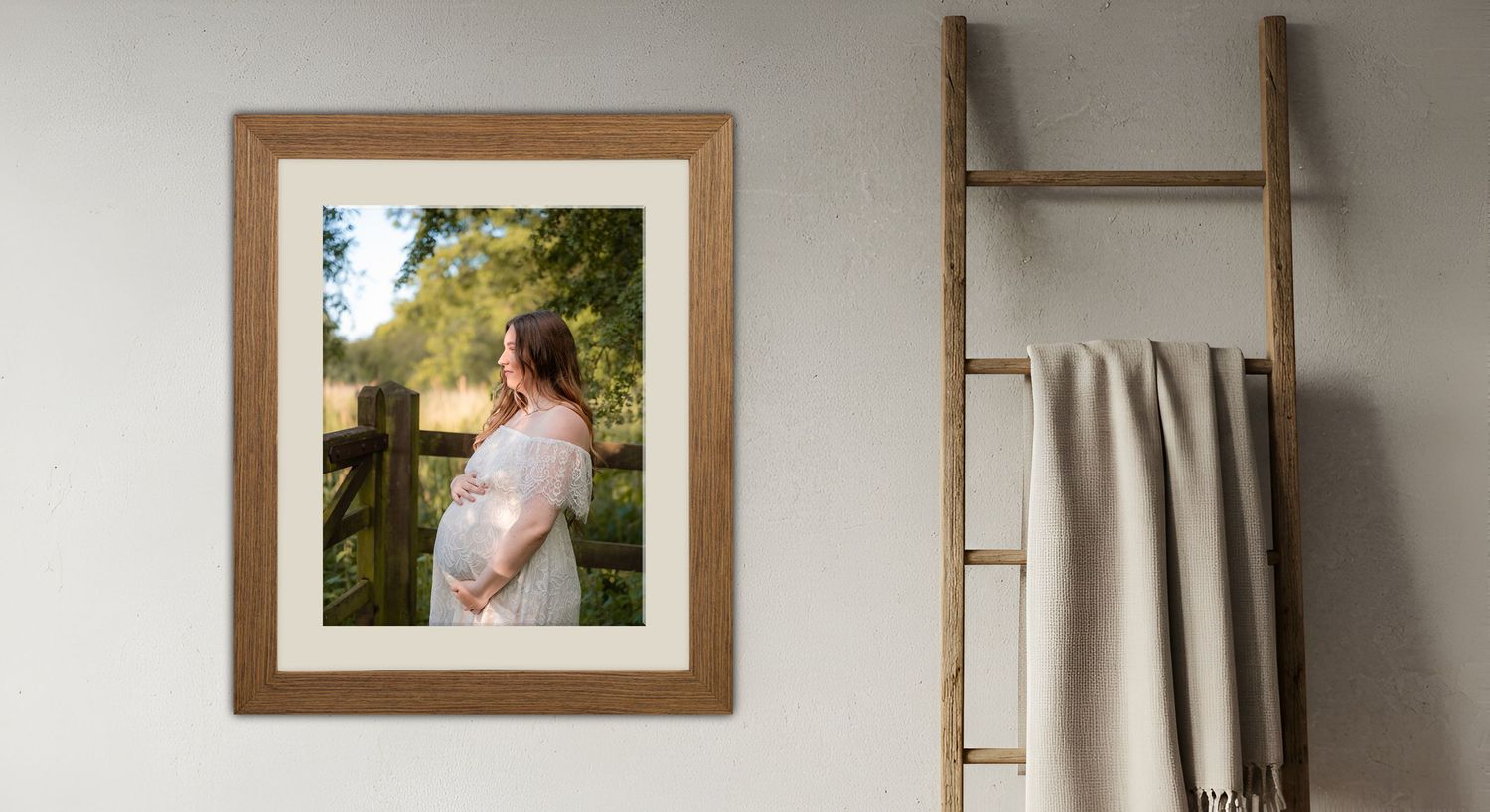 Framed maternity portrait photo in wooden frame displayed on wall beside a rustic wooden ladder with draped towel.