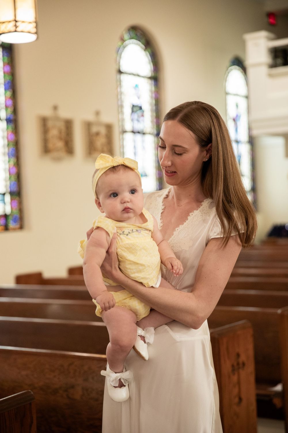 A young woman in a white dress holds a baby girl in a yellow outfit inside a church with stained glass windows.