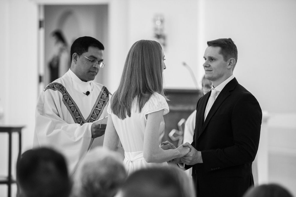 Black and white photo of a couple exchanging wedding vows before a priest in a church ceremony.
