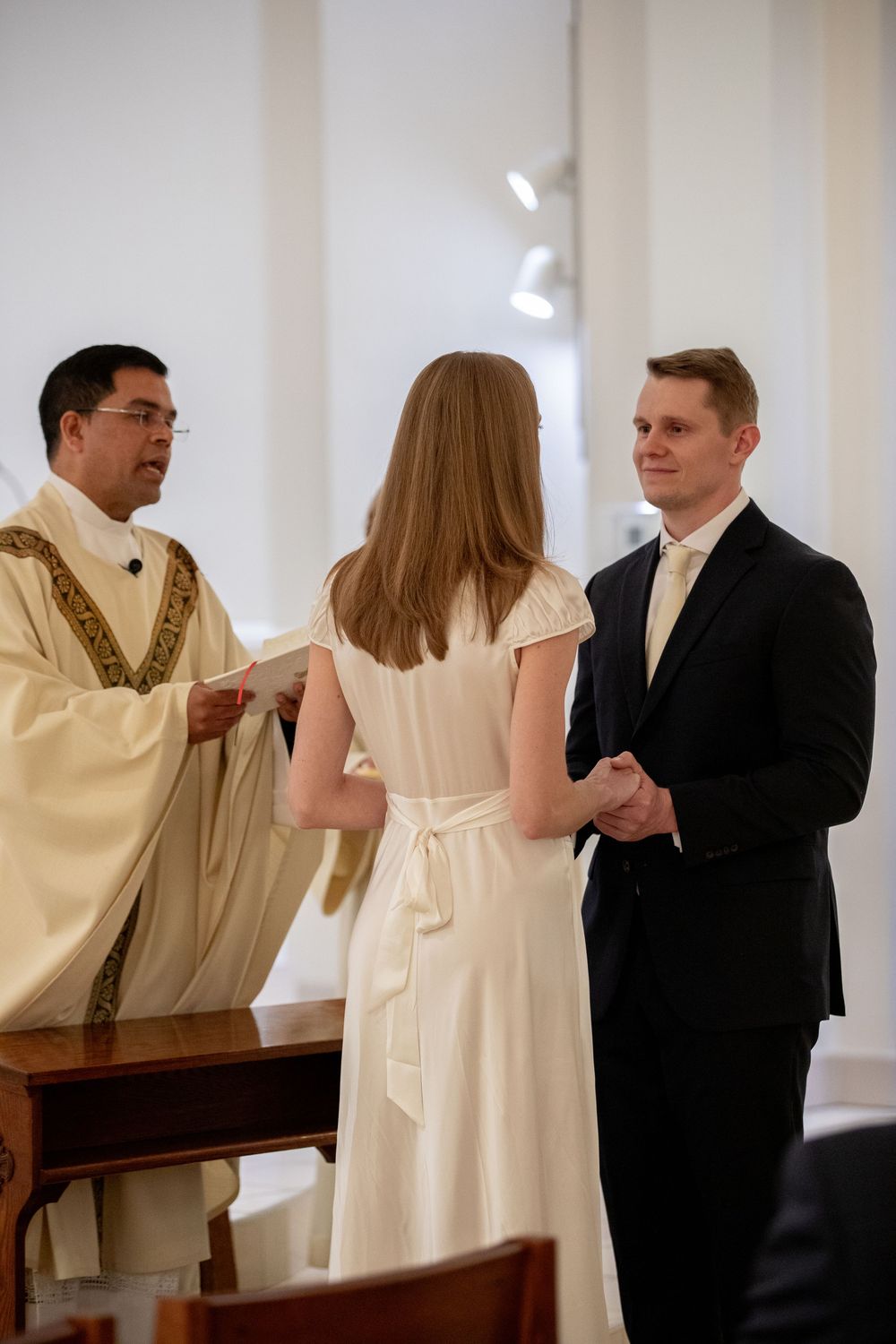 Bride and groom exchange vows before a priest at a Catholic wedding ceremony in a white church.