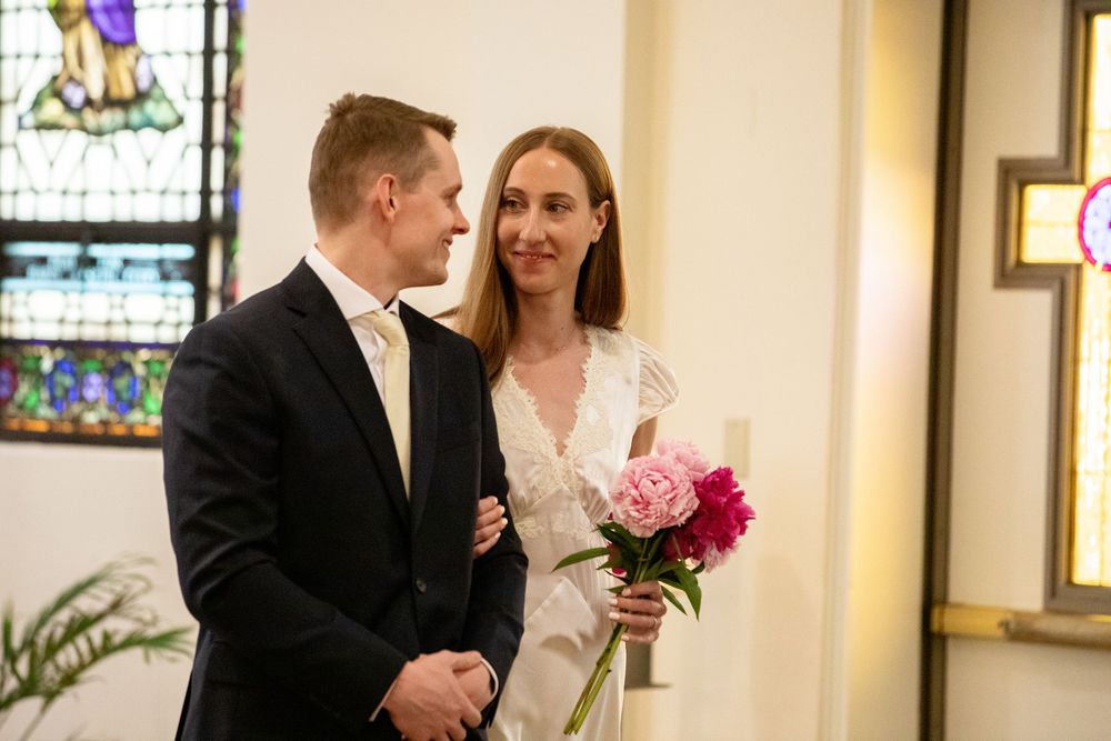 Bride holding pink peonies bouquet stands with groom in navy suit inside church with stained glass windows.