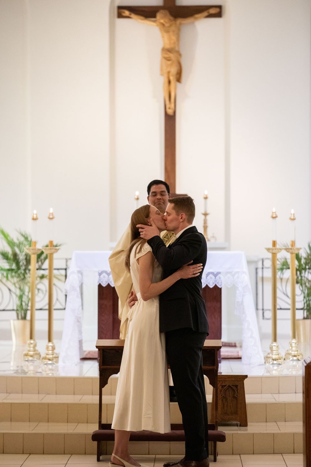 Couple sharing first kiss at Catholic church altar with crucifix backdrop during wedding ceremony.
