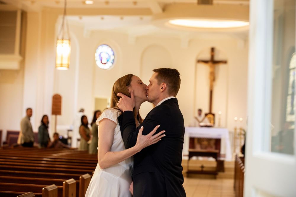 Bride and groom share first kiss at church altar during wedding ceremony with stained glass windows.