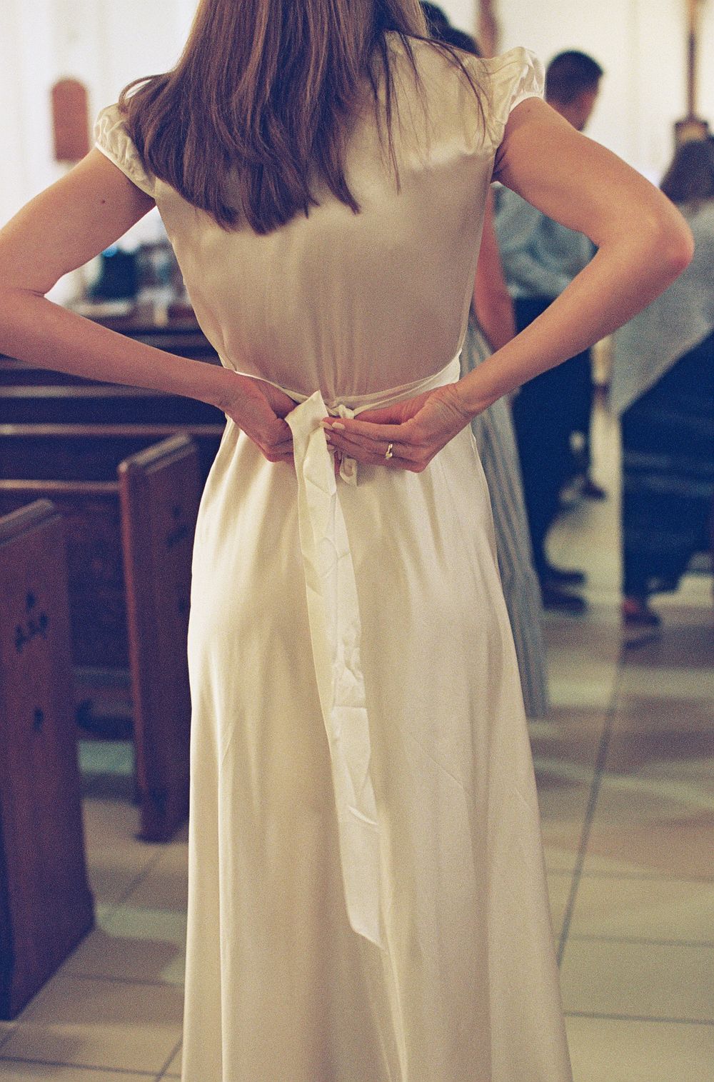 Bride in elegant ivory satin gown tying a bow sash at her waist inside a church.