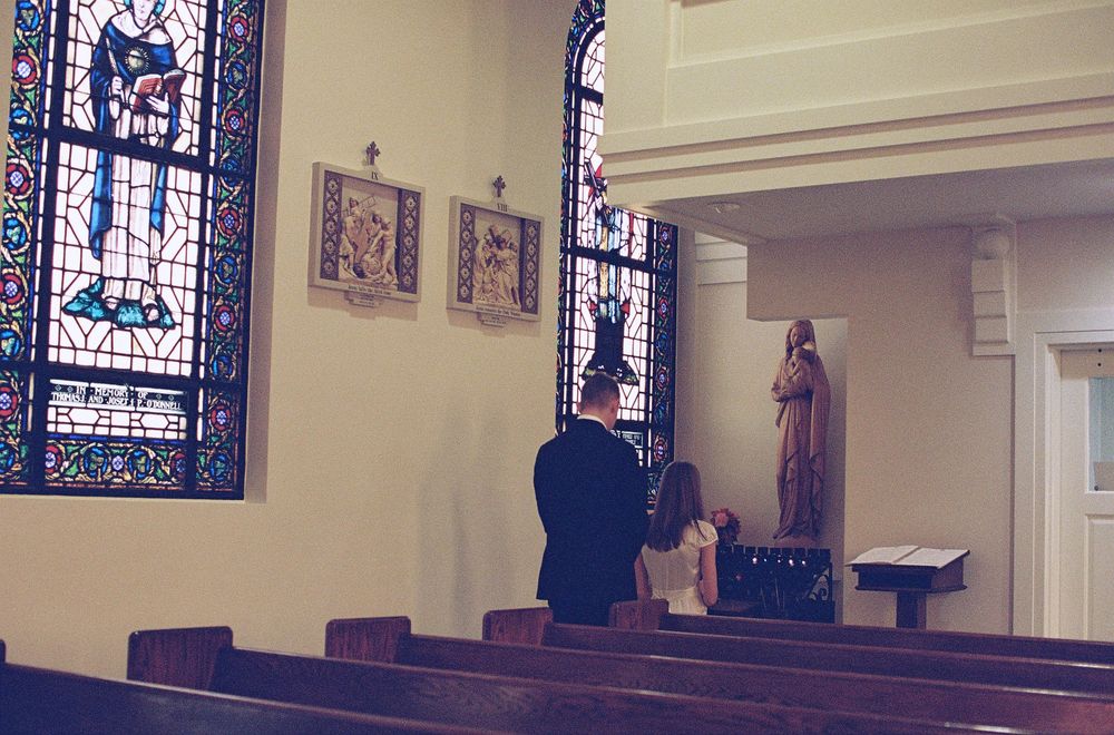 Man and child kneeling in prayer before a statue in a Catholic church with stained glass windows.