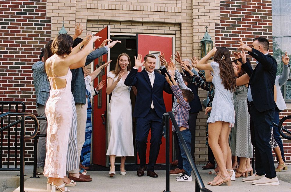 Newlyweds exit through red door as guests form celebratory arch outside brick building.