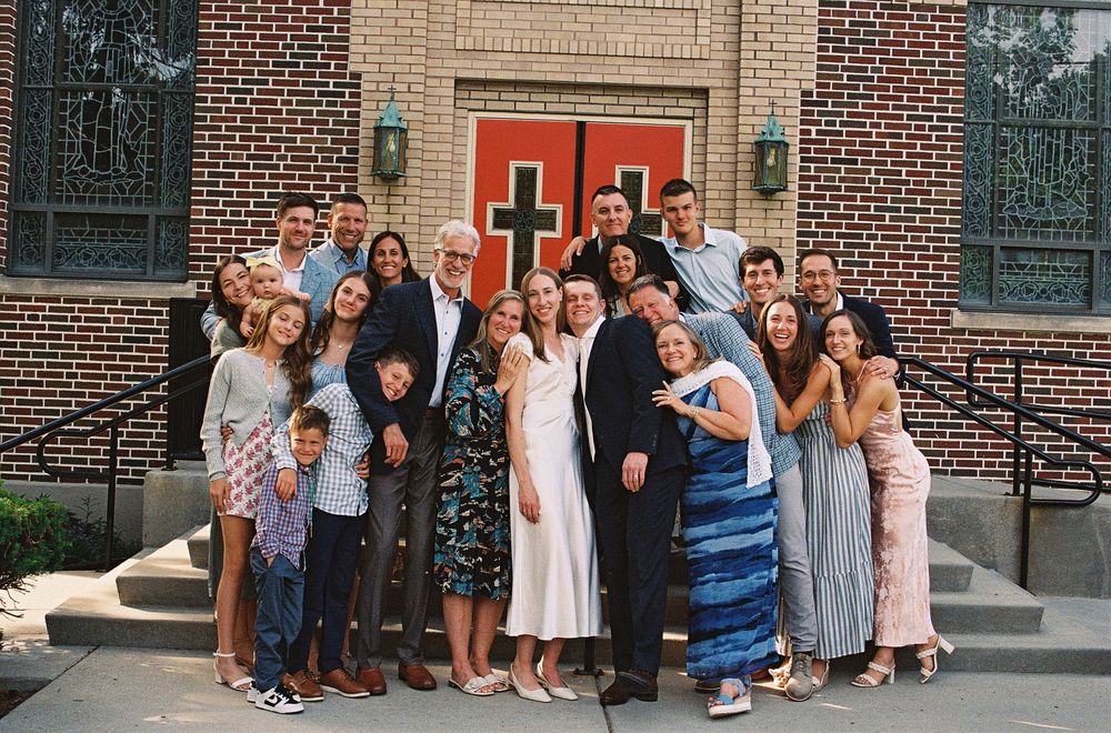 Large family group photo outside a brick church with red doors, celebrating a wedding or special occasion.