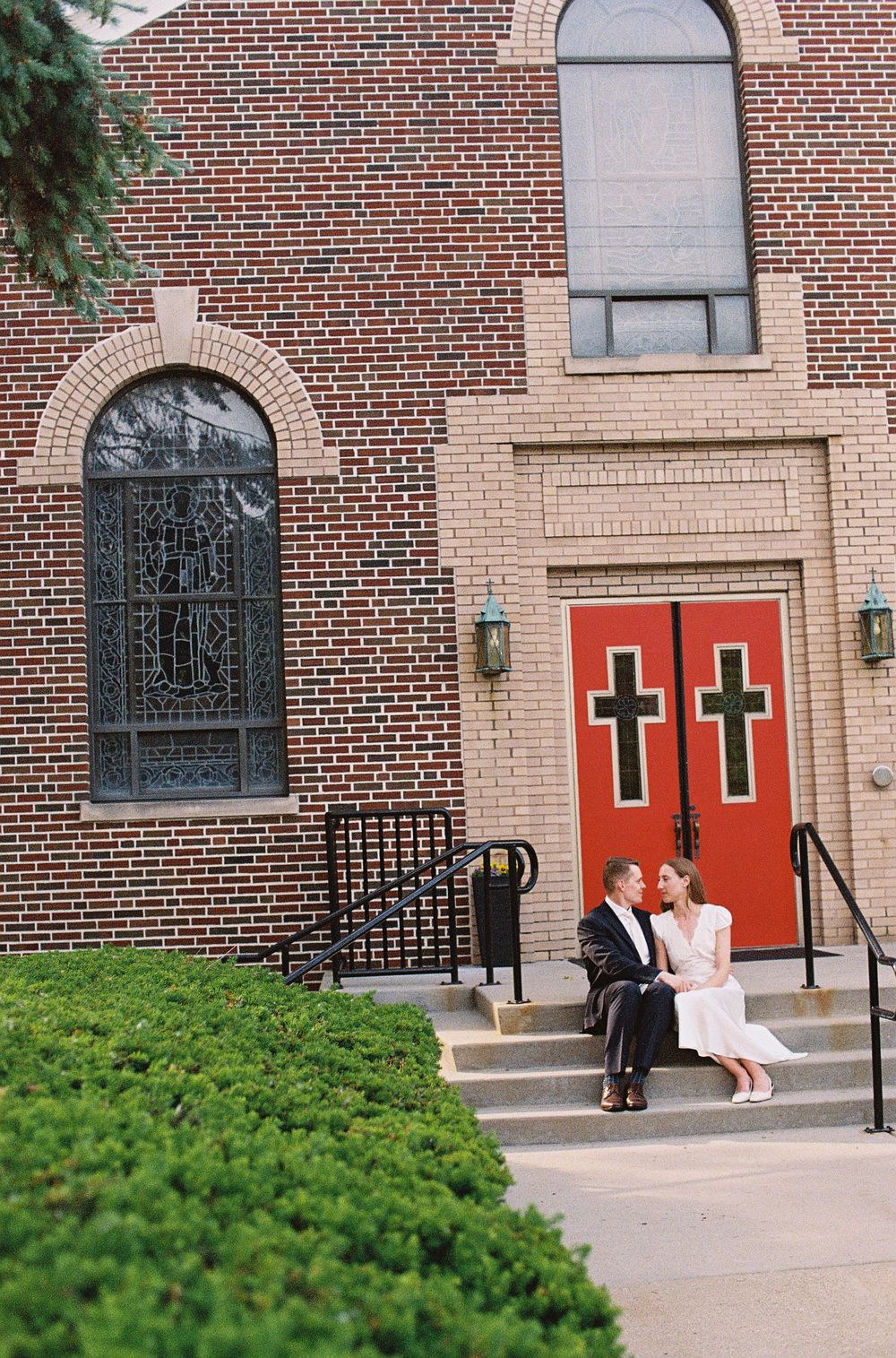 Bride and groom sitting on church steps beside red doors with crosses on their wedding day.