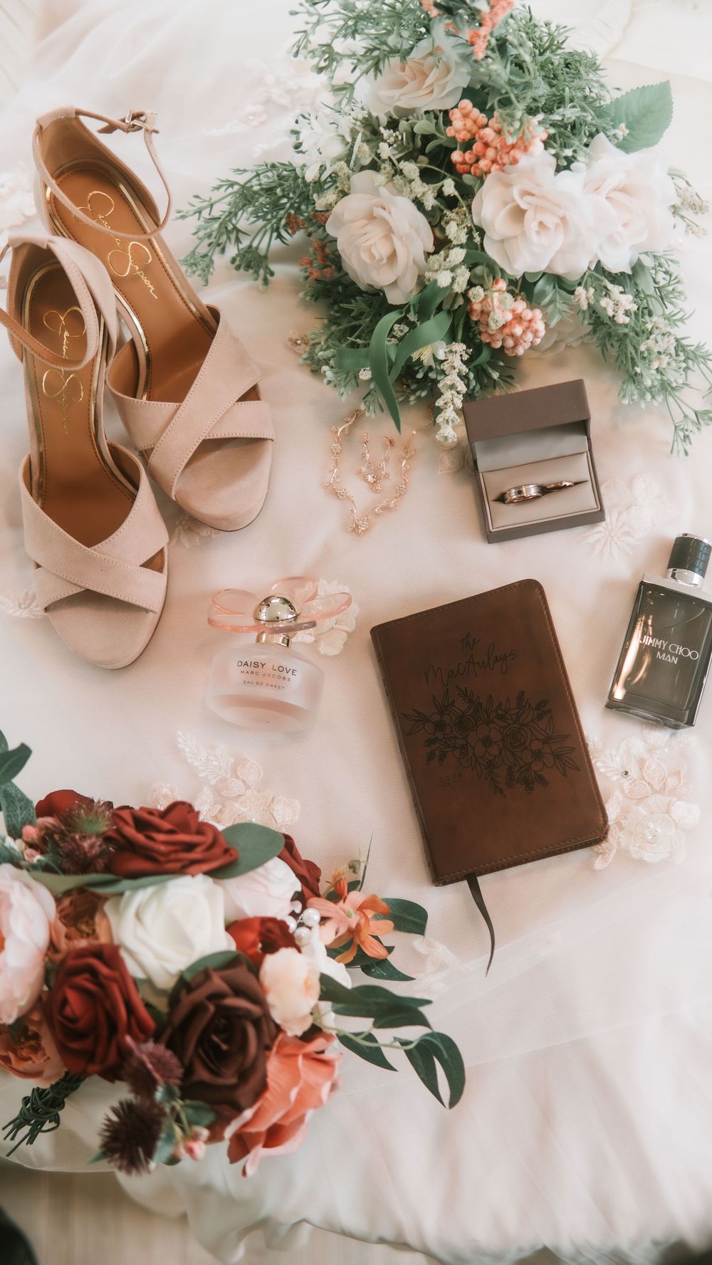 Bridal flat lay featuring nude heels, wedding rings, perfume, a floral journal, and pink and red bouquets on white fabric.