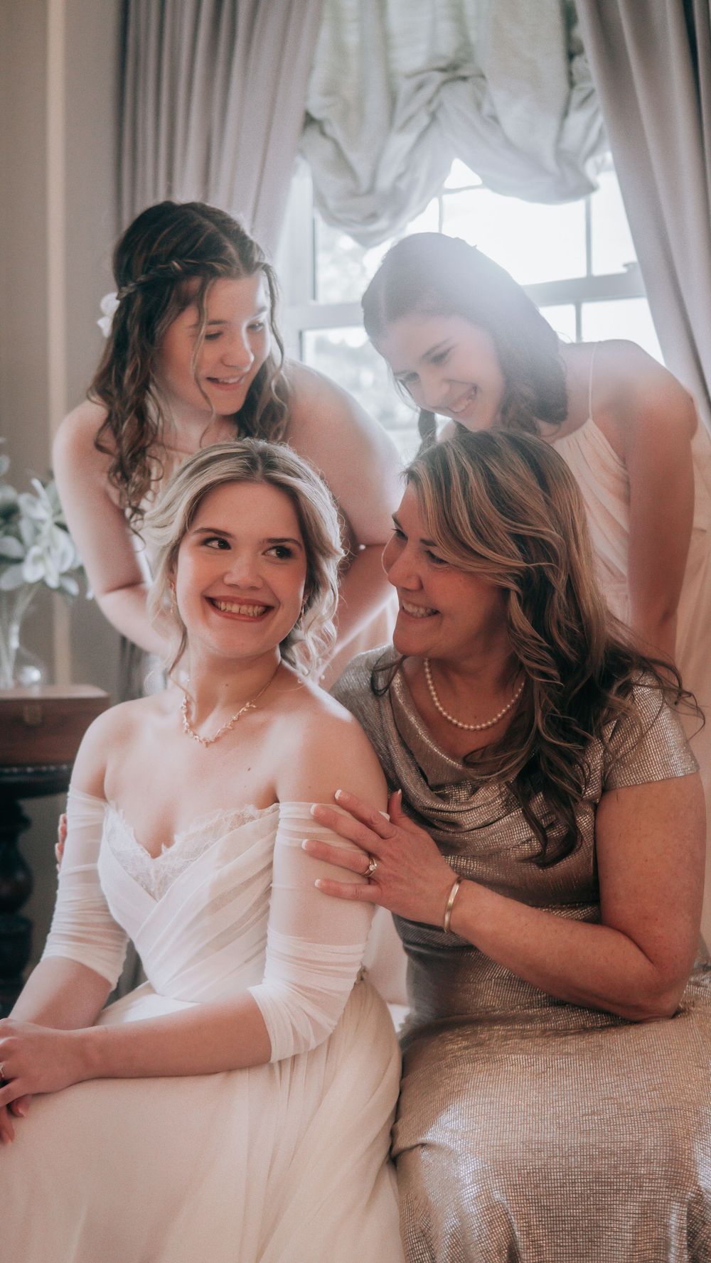 Bride in white gown smiles with mother and two girls in elegant wedding preparation scene indoors.