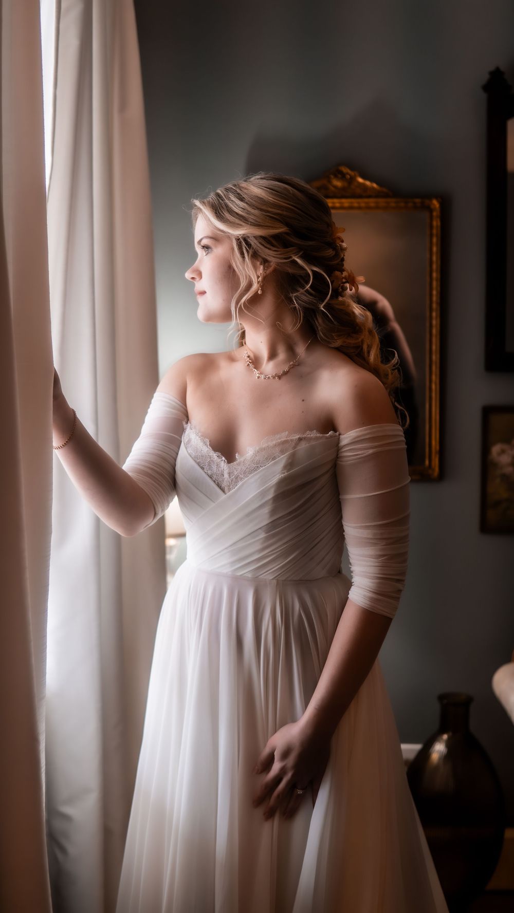 Bride in off-shoulder white gown gazing out window.