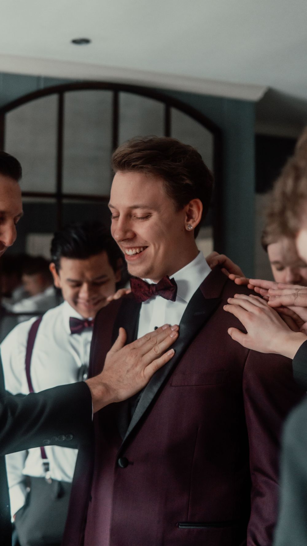 Smiling groom in burgundy tuxedo with bow tie being helped by groomsmen during wedding preparations.