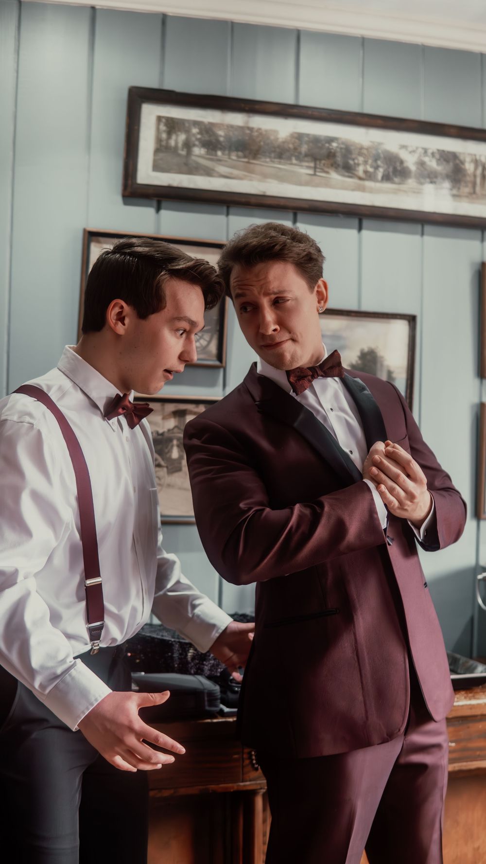 Two grooms in burgundy bow ties prepare for their wedding, one helping the other with his suit jacket.