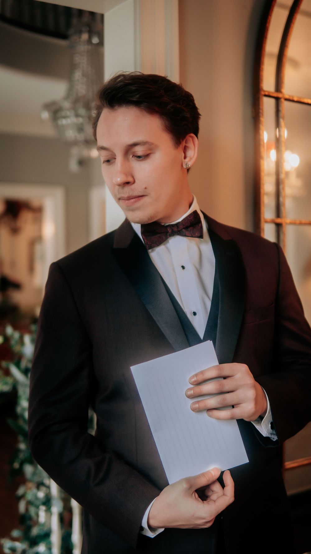 Elegant groom in black tuxedo with burgundy bow tie reading a card indoors before the wedding ceremony.