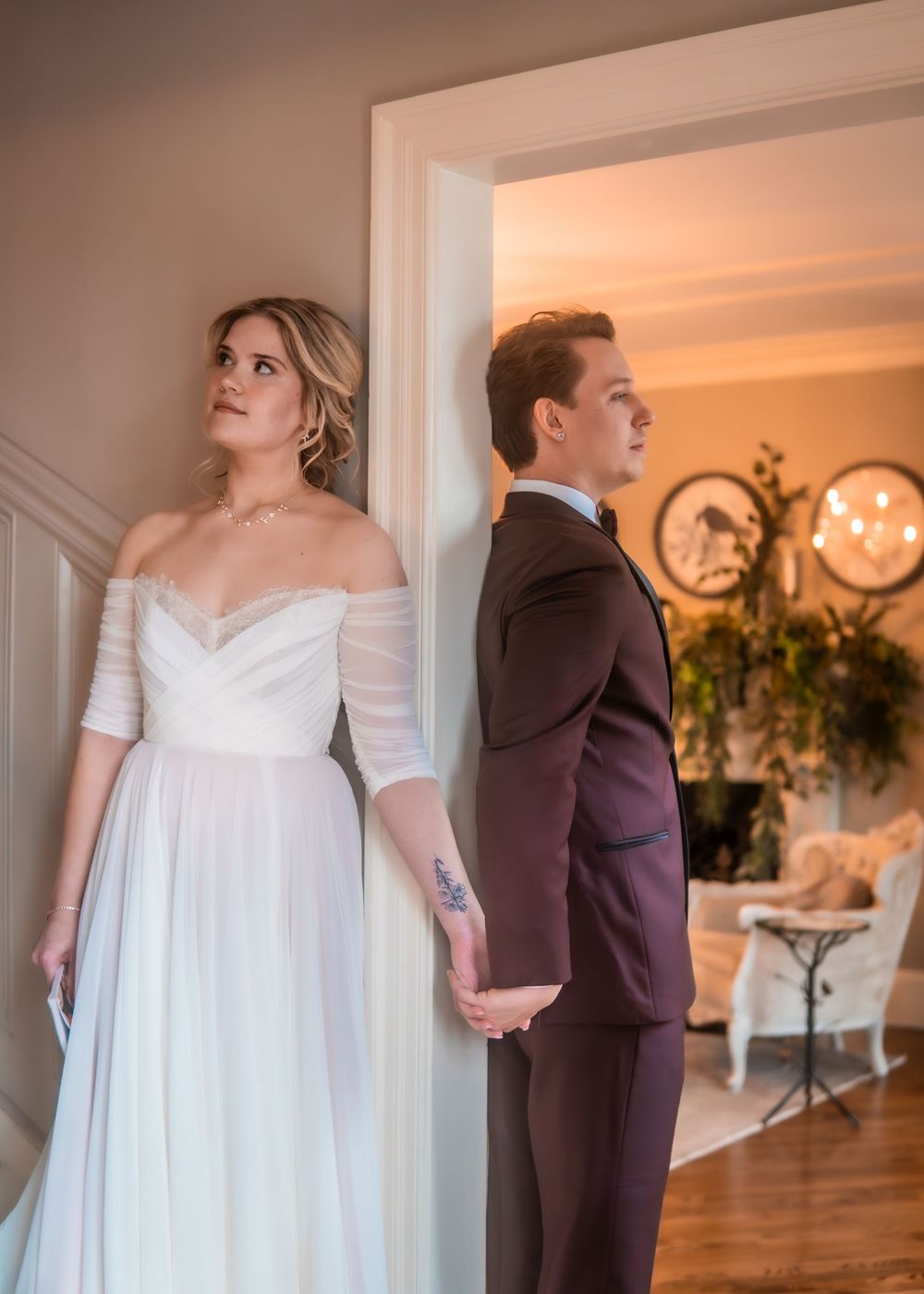 Bride in white off-shoulder gown and groom in burgundy suit hold hands in elegant doorway during wedding day.