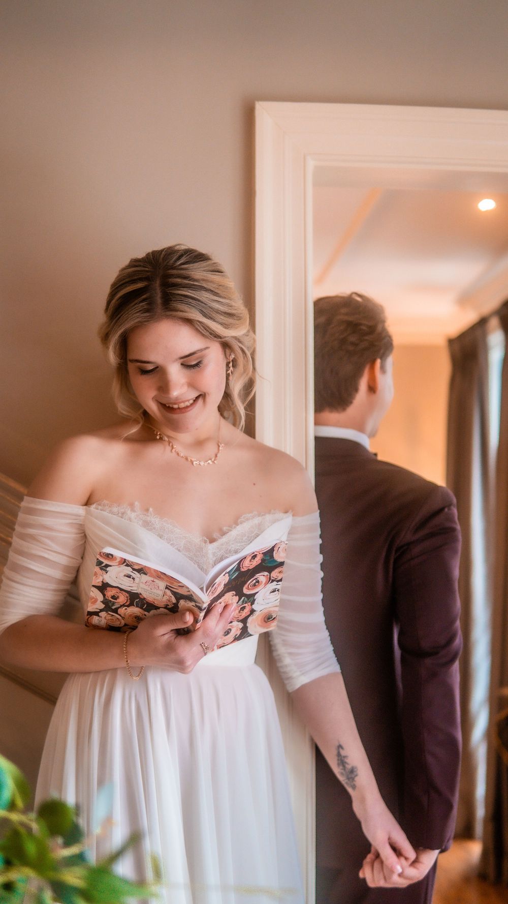 Bride in off-shoulder white gown reads vows from floral book while groom in dark suit holds her hand indoors.