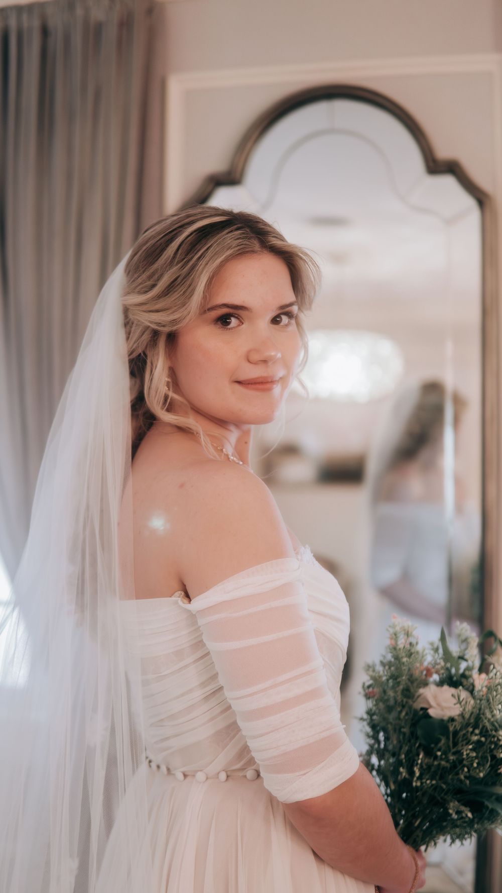 Elegant bride in an off-shoulder white gown and veil holds a bouquet before her wedding.