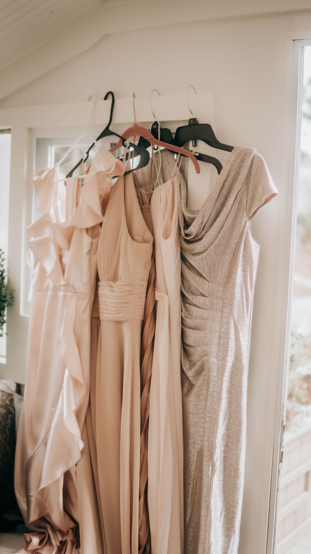 Four elegant blush and champagne bridesmaid dresses hanging on hangers in a bright, airy room.