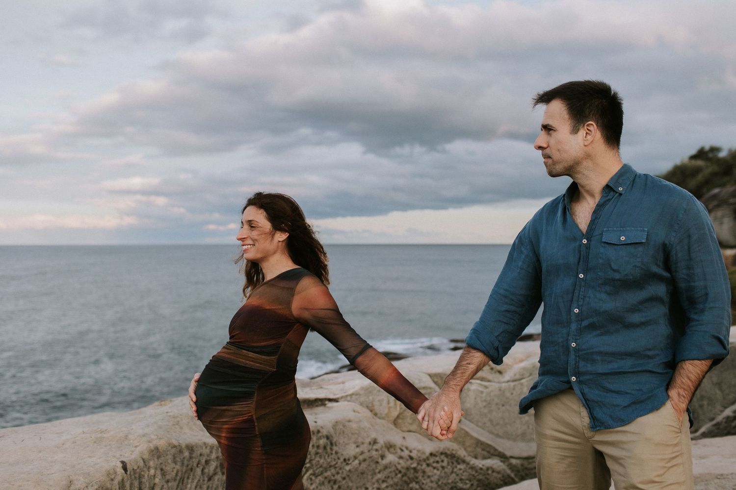 Pregnant couple holding hands on rocky shoreline with ocean and cloudy sky backdrop during maternity photo session.