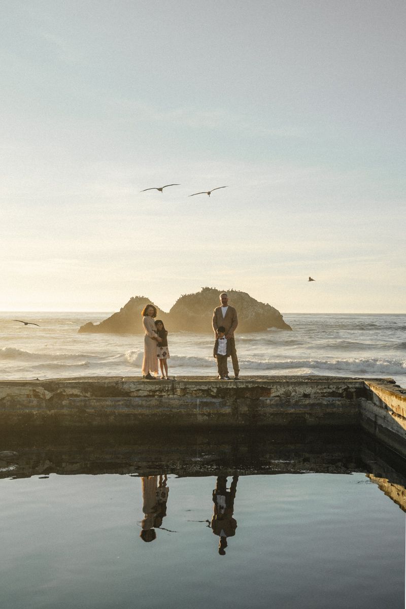 Couple standing on coastal ruins at sunset with ocean waves, rocky islands, seagulls, and a still water reflection below.
