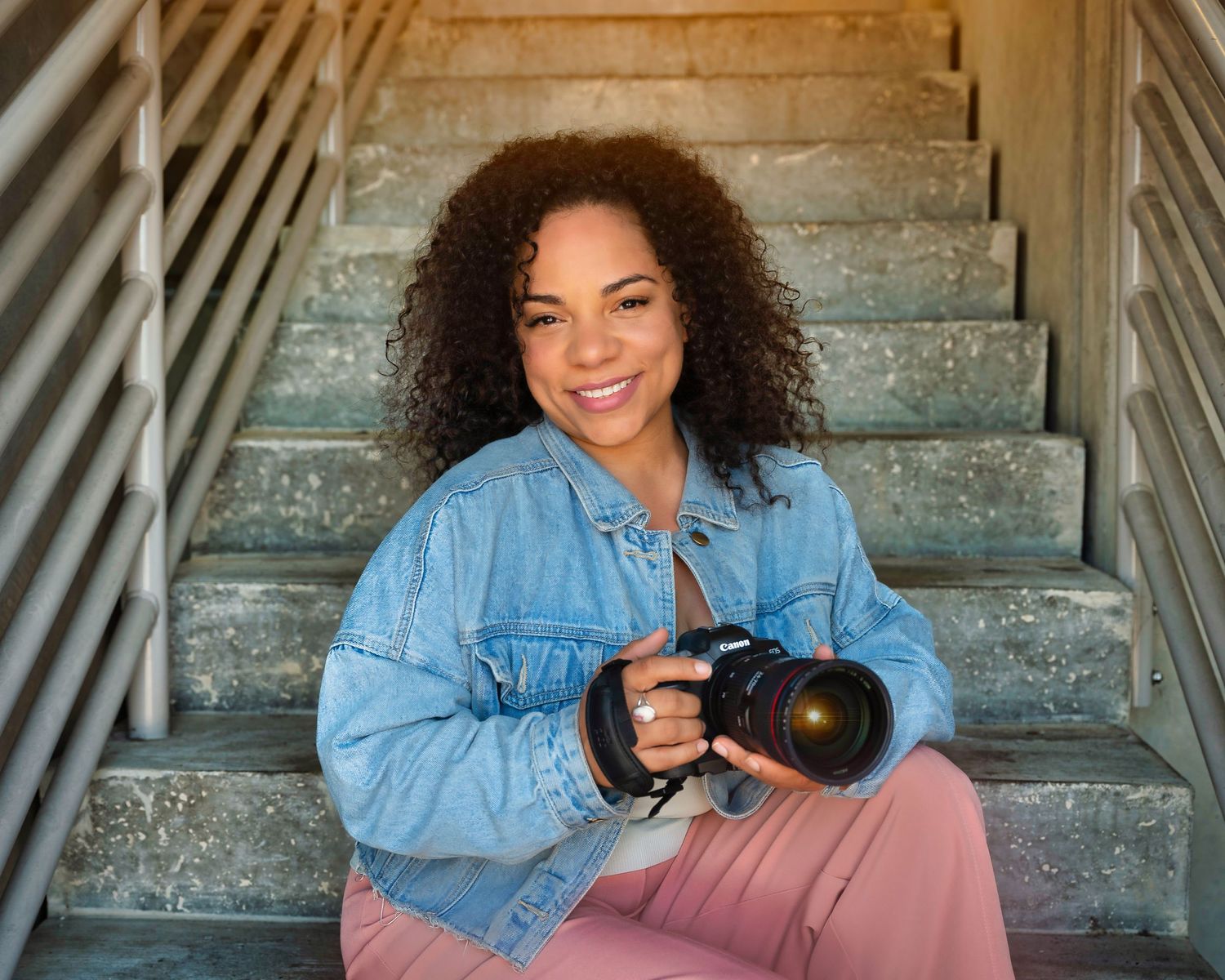 Professional photographer in denim jacket sitting on concrete stairs with camera.