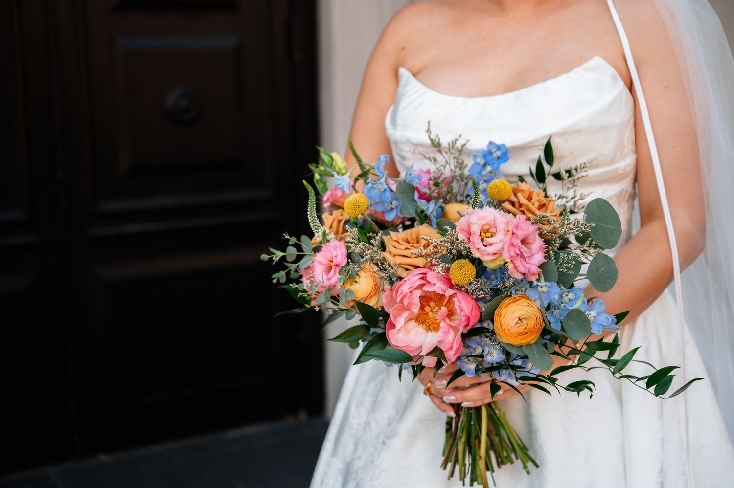 Bride holding a vibrant wedding bouquet with pink peonies, orange roses, blue flowers, and eucalyptus greenery.