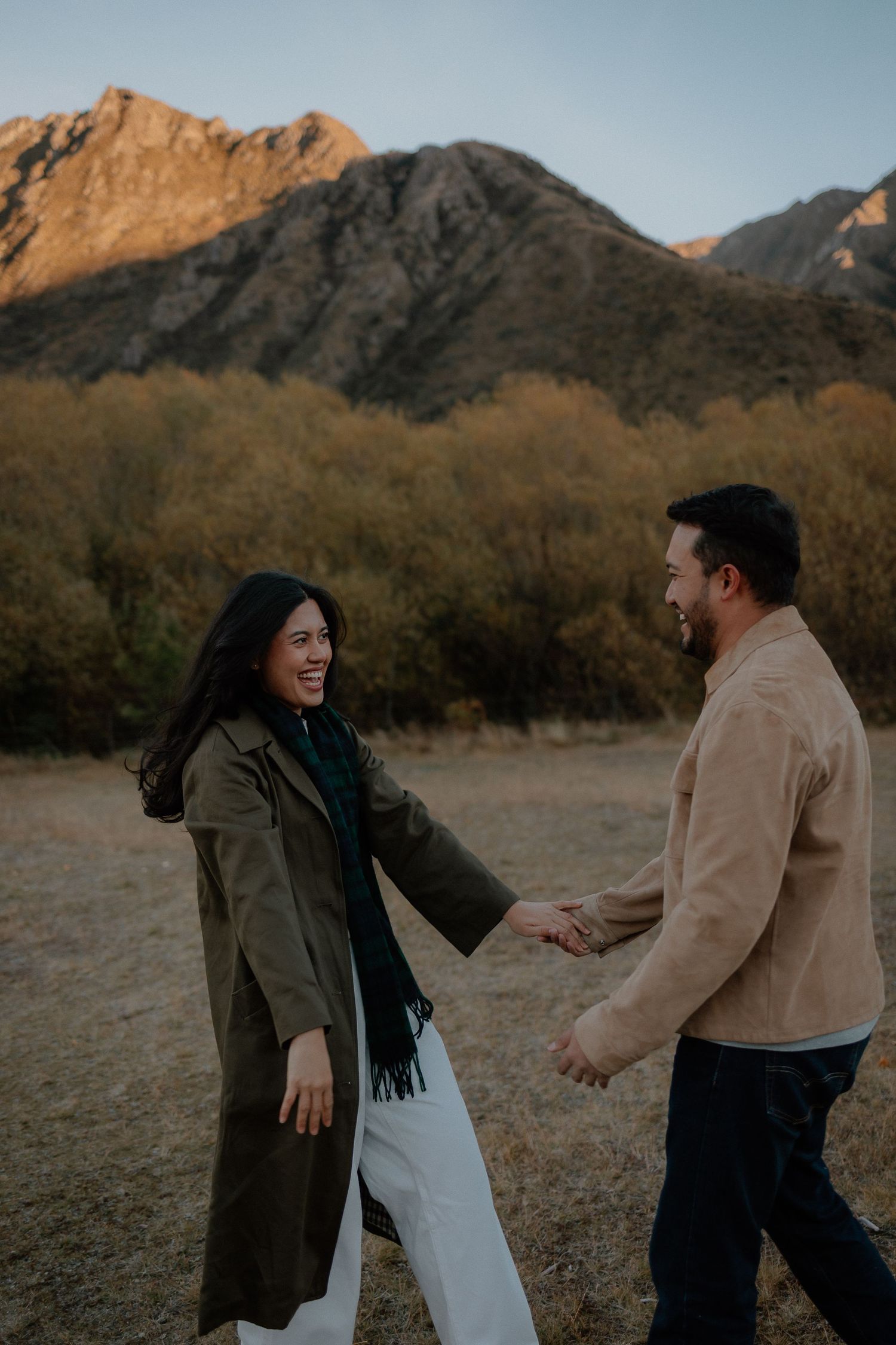 A joyful couple holds hands and laughs together in a mountain meadow at dusk, dressed in autumn outerwear.