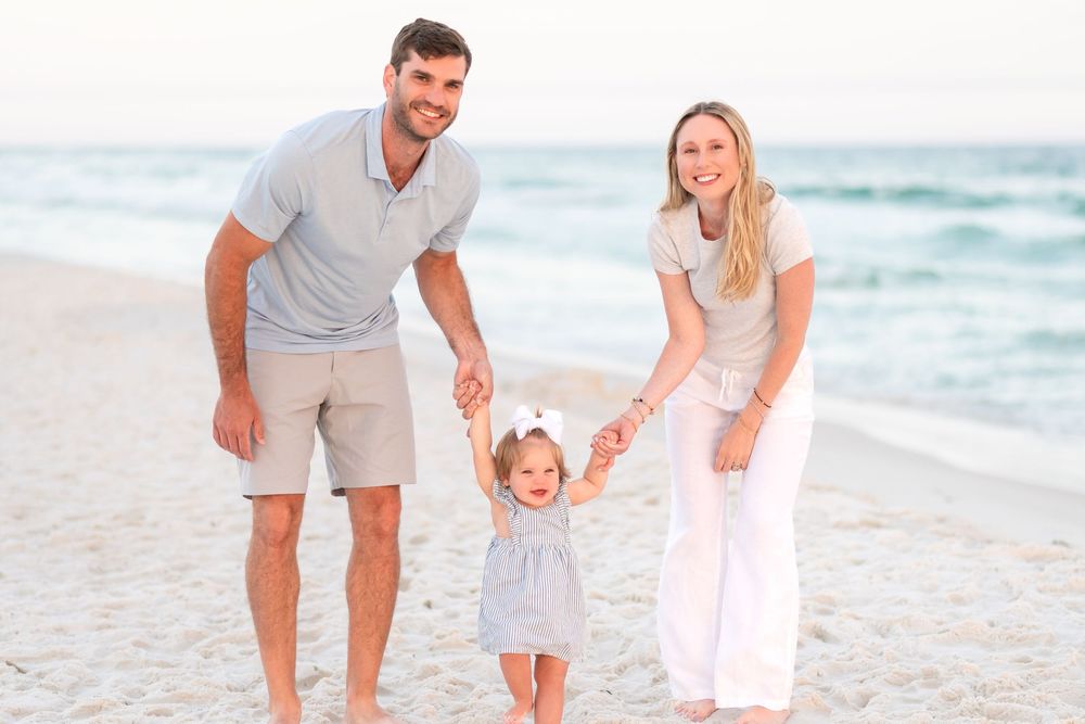 Happy family of three walking on a sunny beach, parents holding toddler girl's hands near ocean waves.