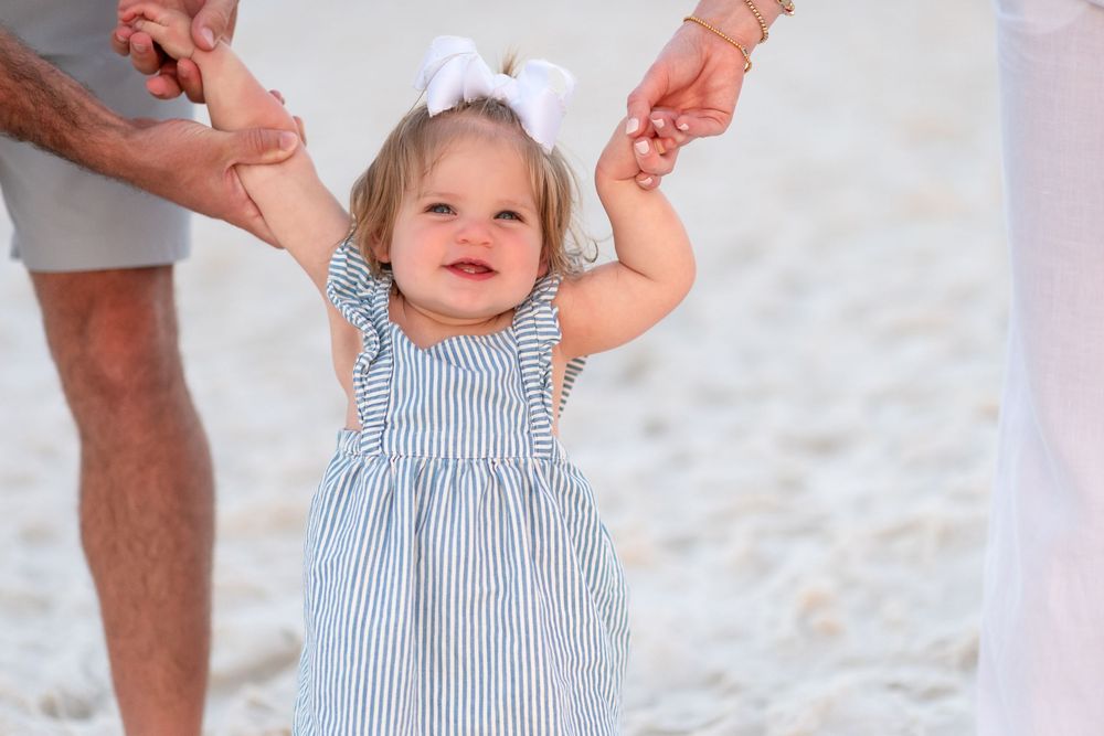 Smiling toddler girl in striped dress with white bow being held by parents on a sunny beach.