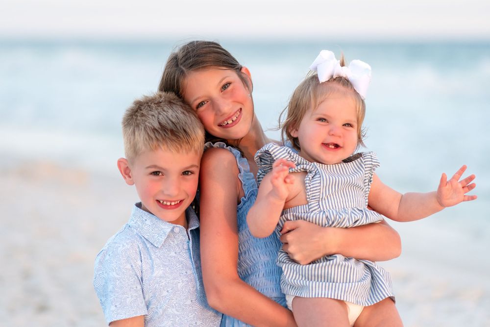 Three happy children pose together on a sunny beach, with the oldest girl holding a smiling toddler in a bow.