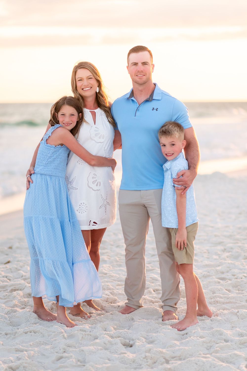 A happy family of four poses together on a sandy beach at sunset, wearing coordinating blue and white outfits.