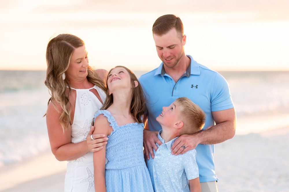 Family of four smiling and laughing together on a sandy beach at sunset, wearing coordinating blue and white outfits.