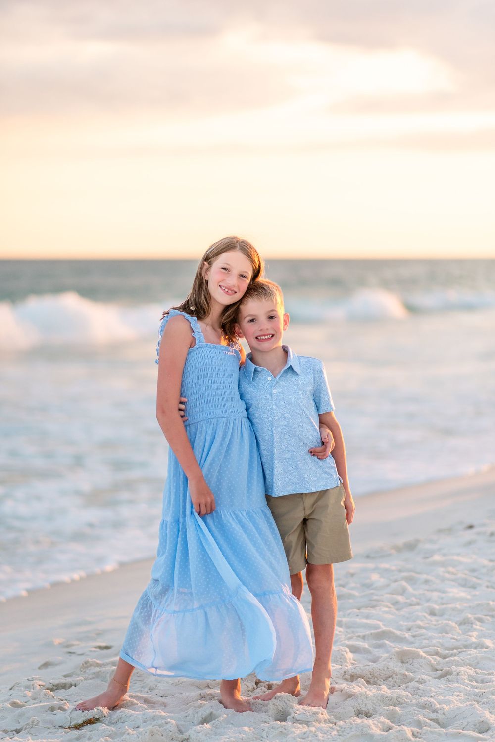 Two smiling children in blue outfits pose together on a sandy beach at sunset with ocean waves behind them.