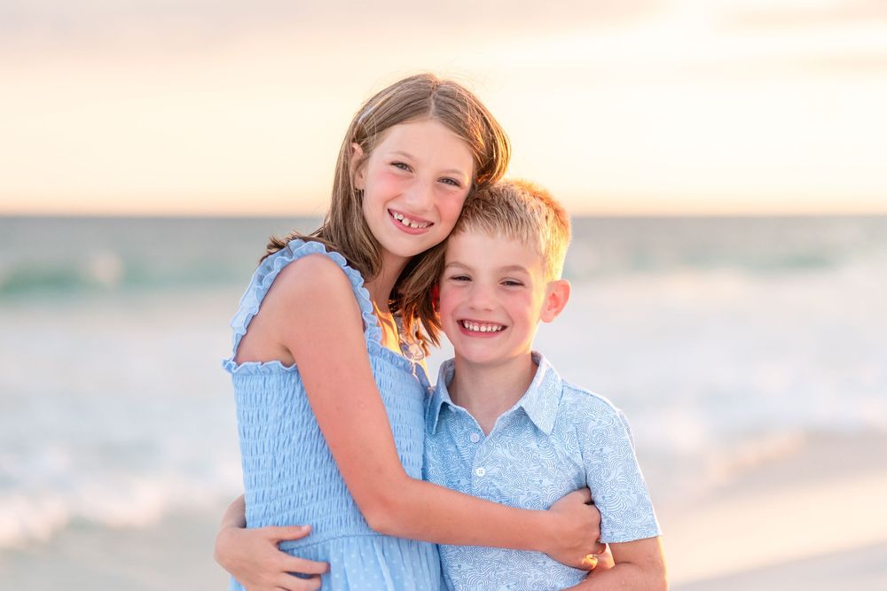 Two smiling children hugging on a sandy beach at sunset, wearing blue outfits with ocean waves in the background.