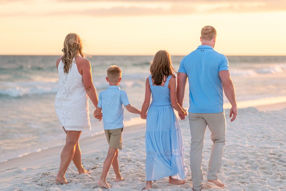 Family of four holding hands walking toward ocean waves at golden sunset on a sandy beach.