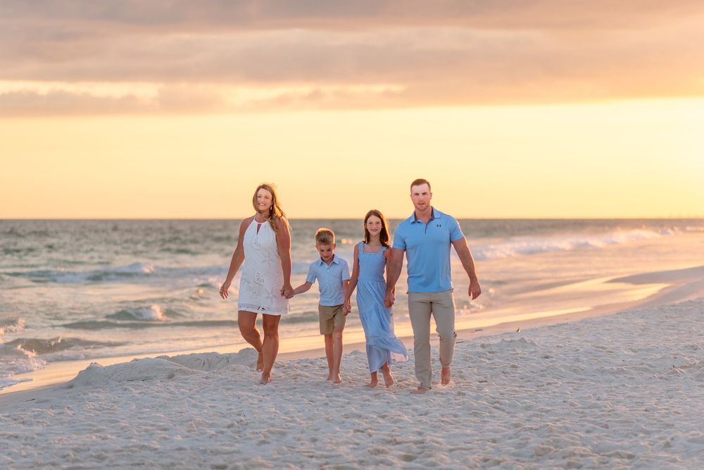 Family of four walking barefoot on sandy beach at golden sunset, wearing white and blue outfits, waves in background.
