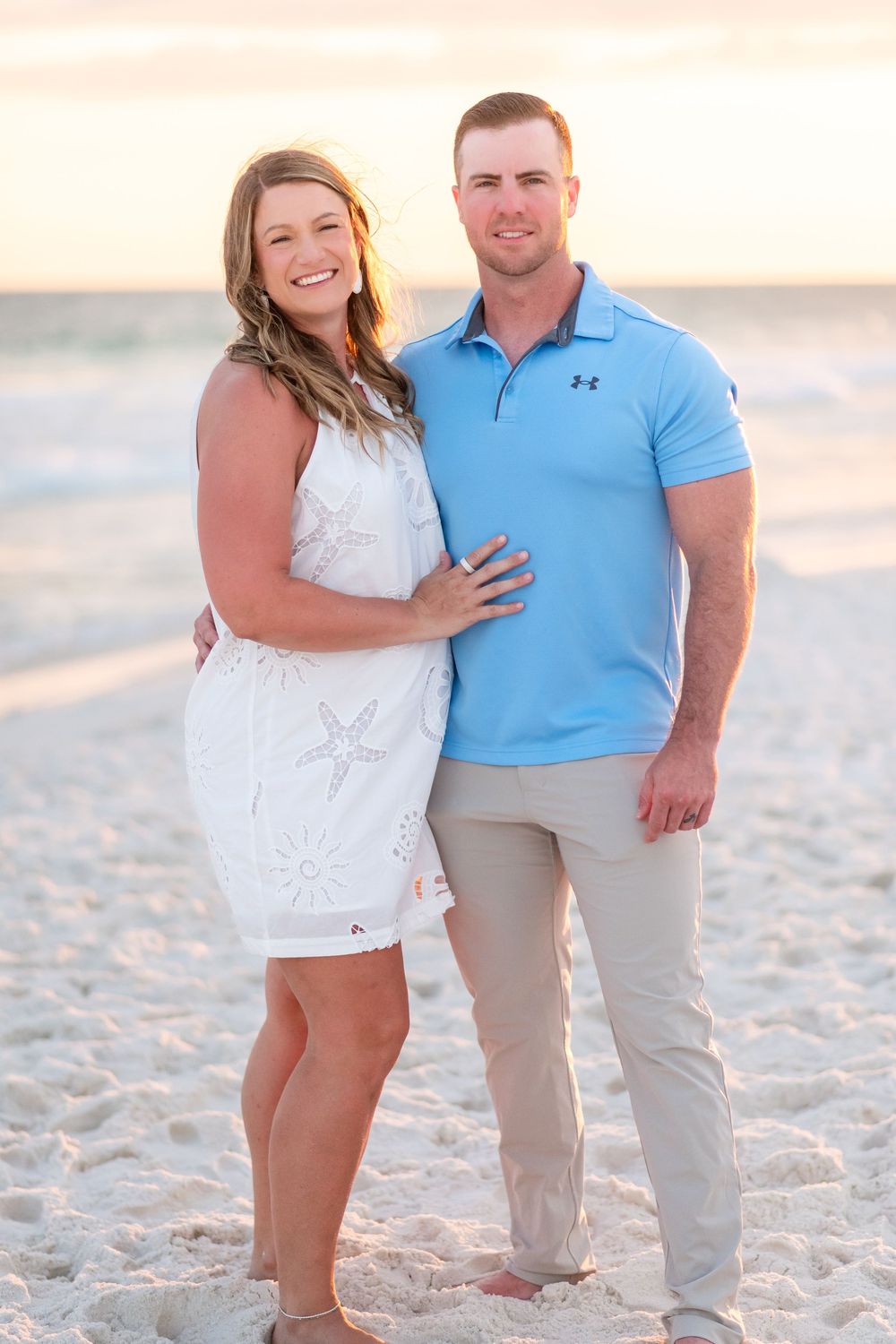Smiling couple posing together on a sandy beach at sunset, woman in white dress and man in blue polo shirt.