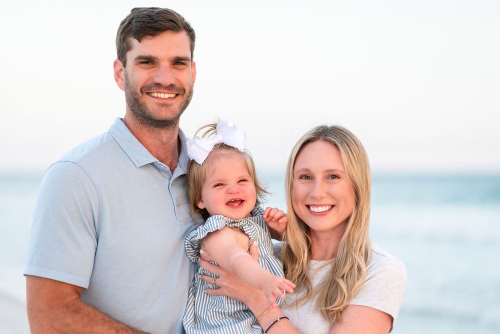 A smiling family of three poses on a beach, with parents holding their happy baby girl wearing a white bow.