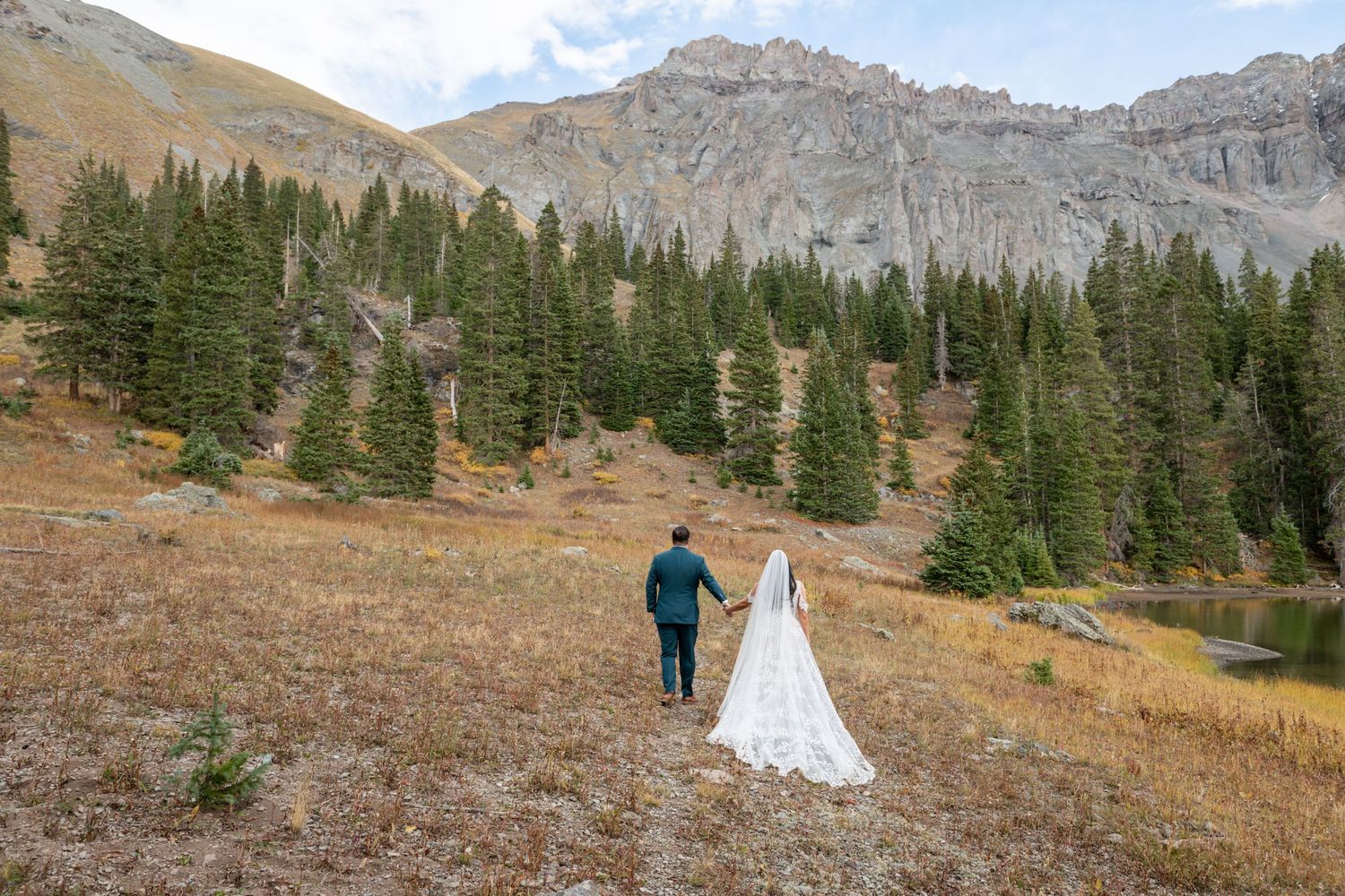 Bride and groom holding hands walking through a mountain meadow near Telluride with pine trees and rocky peaks in background.