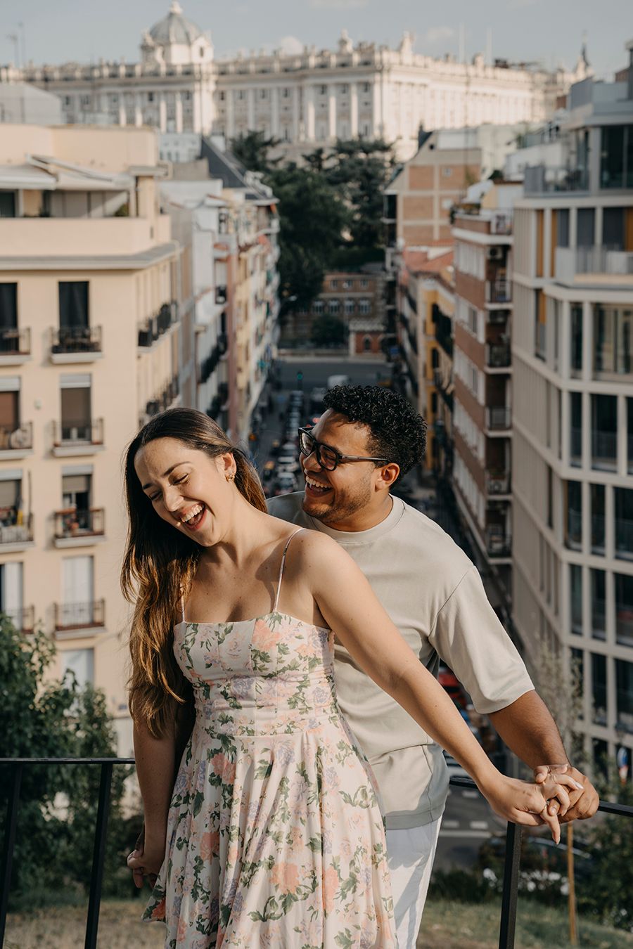 Happy couple holding hands on a balcony overlooking a European city street with historic buildings.