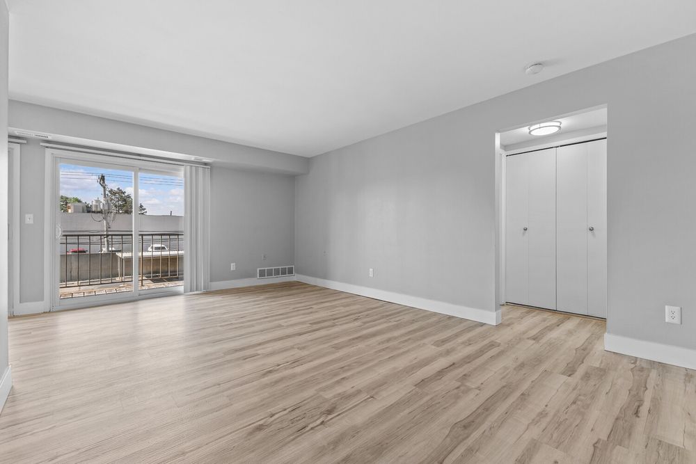 Bright empty living room with light wood floors, sliding glass door to balcony, and white closet doors.
