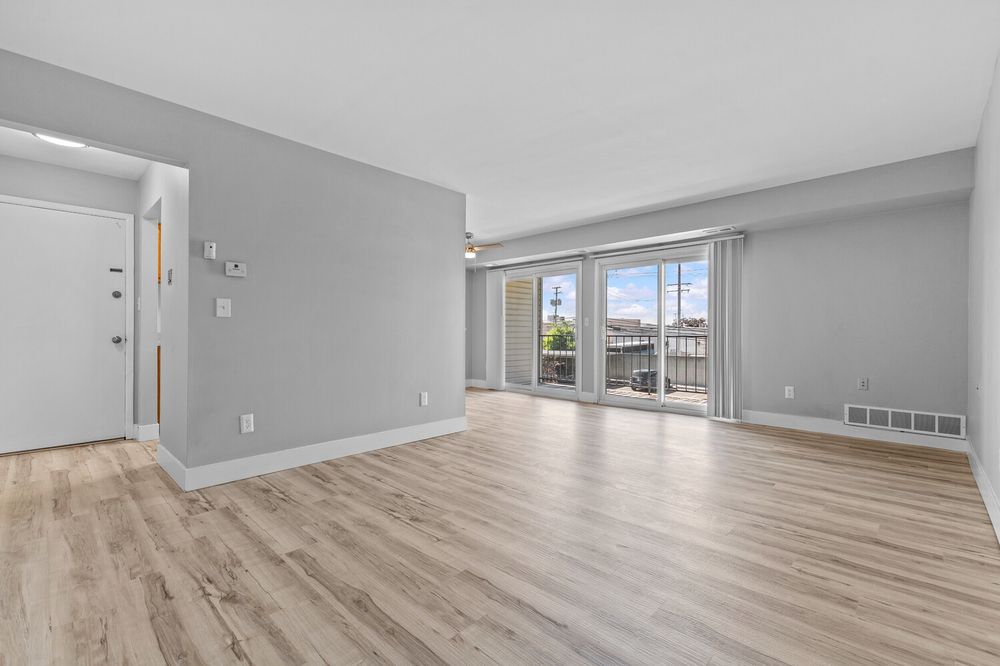 Bright open living room with light hardwood floors, gray walls, white door, and sliding glass doors leading to a balcony.